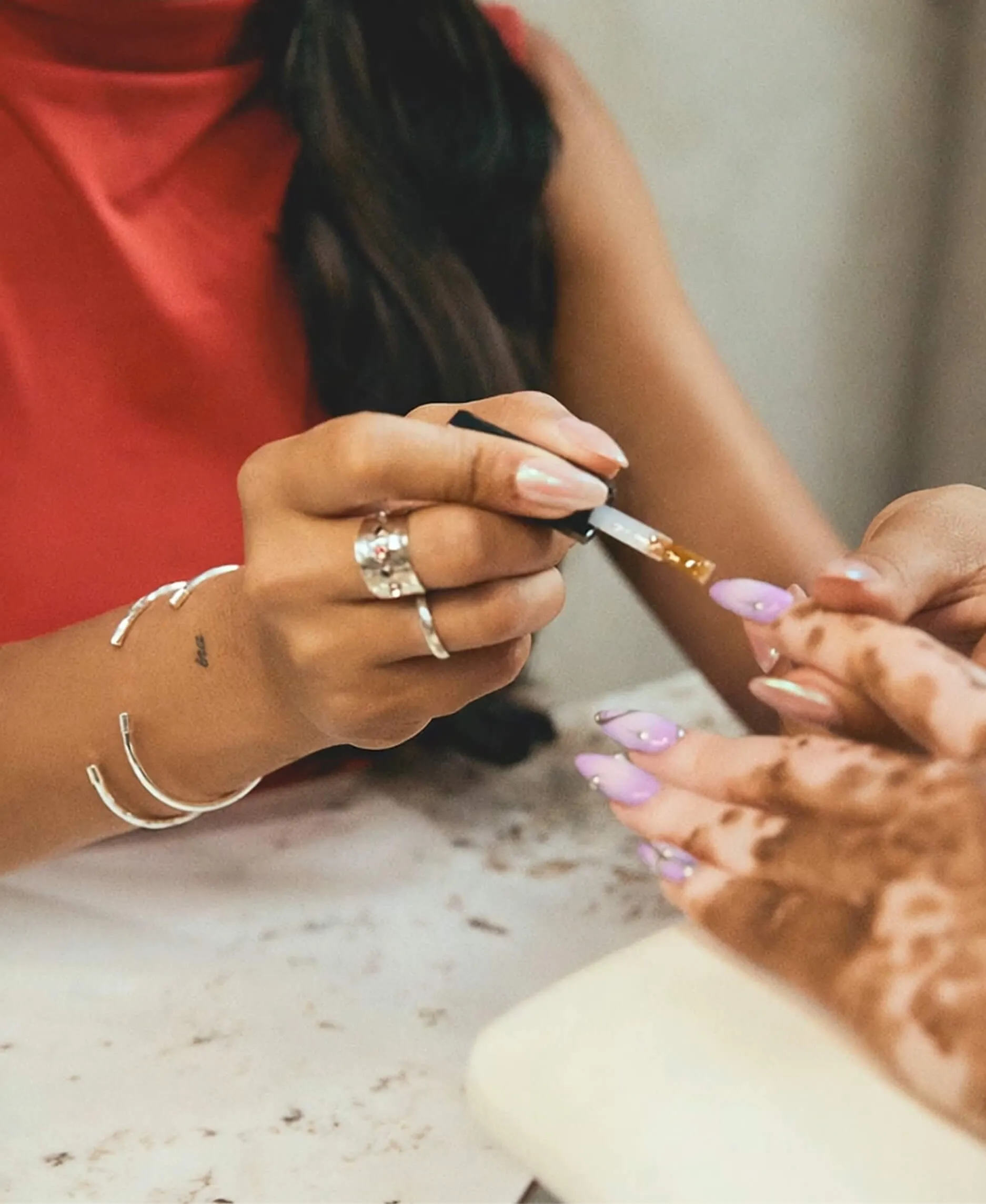 Image of a nail tech with lots of jewlery giving a pink manicure to a customer.